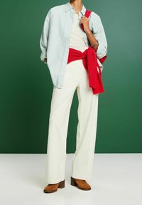 Chemise oversize bleu clair rayée, pantalon large blanc, pull rouge noué à la taille, et chaussures à talons marron. Fond vert uni.