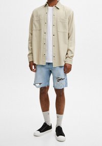 Beige-kauluspaita, jossa on kaksi rintataskua, valkoisen t-paidan päällä. Vaaleansiniset repaleiset farkkushortsit. Mustat lenkkitossut, joissa on valkoiset pohjat.