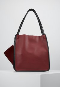 Borsa a mano in pelle bordeaux con manici e rifiniture nere, dotata di una pochette staccabile coordinata, esposta su un piedistallo bianco.