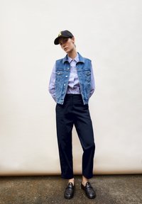 Gilet in denim sopra una camicia a righe con bottoni, abbinato a pantaloni ampi blu navy e scarpe slip-on nere, completato da un cappellino da baseball nero.