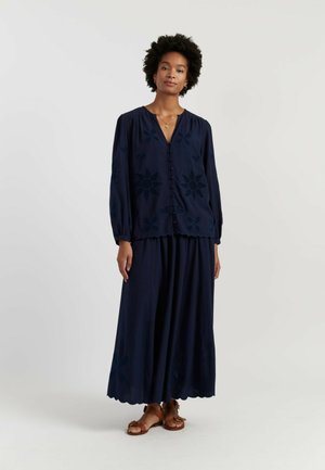 Femme portant un chemisier bleu marine à manches longues et un pantalon large assorti avec des découpes fleurs, associé à des sandales marron.