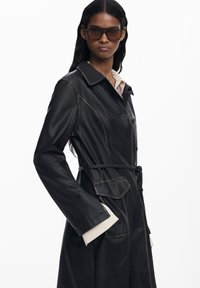 Cappotto trench in pelle nera con cuciture a vista, dotato di una vita con cintura, maniche larghe e tasche laterali. Il modello indossa occhiali da sole scuri.