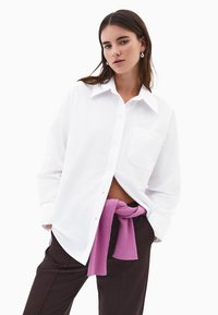 Hvid langærmet skjorte med klassisk krave og brystlomme, parret med mørkebrune bukser og en lyserød striktrøje bundet om livet.