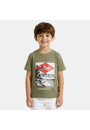 Jeune garçon souriant portant un t-shirt graphique vert olive avec un motif de plage et un pantalon blanc, debout devant un fond uni.