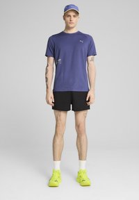 Violeta Puma t-krekls ar logo, melnas šortus un neona zaļus sporta apavus. Cepure saskan ar kreklu un tai ir dzeltena akcenta krāsa. Redzamas tetovējumi uz rokām.