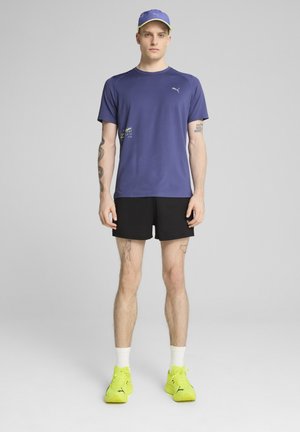 Fioletowa koszulka Puma z logo, czarne spodenki i neonowozielone sneakersy. Czapka pasuje do koszulki z żółtym akcentem. Tatuaże widoczne na ramionach.