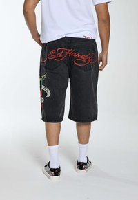 Shorts di jeans neri con il logo cursivo rosso "Ed Hardy" sul retro, caratterizzati da grafiche multicolori e una texture leggermente sbiadita.