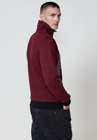 Suéter de manga larga texturizado en rojo con cuello alto y detalles en negro, que presenta un estampado gráfico en la parte delantera y puños y dobladillo acanalados.