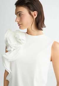 Top bianco senza maniche con un'accenno di rouches testurizzato sulla spalla sinistra, realizzato in tessuto leggero e con una texture liscia.