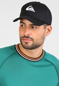 Svart caps med en hvit logo, kombinert med en grønn hettegenser med stripete krage i svart, hvitt og oransje.