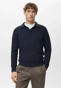 Marineblauwe gebreide trui met een geribbelde textuur, voorzien van een V-hals en een contrasterende kraag. Gecombineerd met lichtbruine broek.