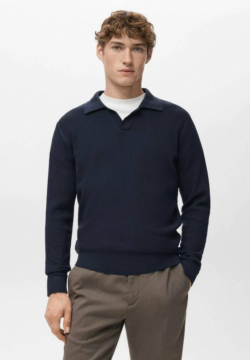 Marineblauwe gebreide trui met een geribbelde textuur, voorzien van een V-hals en een contrasterende kraag. Gecombineerd met lichtbruine broek.