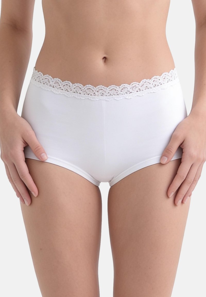Femme portant un shorty en coton blanc bordé de dentelle, visible de la taille jusqu’en haut des cuisses sur un fond uni.