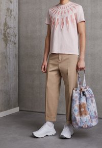 Světle růžové tričko s potiskem blesků, béžové zkrácené kalhoty a bílé tenisky. Drží tašku s tie-dye vzorem v modré a hnědé barvě.