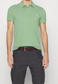 Grön polo skjorta med krage, med en rosa logotyp. Bärs tillsammans med mörkgrå cargobyxor och ett brunt bälte, med flera fickor.