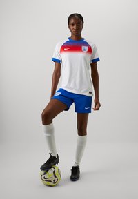 Camiseta blanca de fútbol para mujer con cuello en degradado rojo y detalles azules, combinada con pantalones cortos azules, botas negras y calcetines blancos, de pie sobre un balón de fútbol.