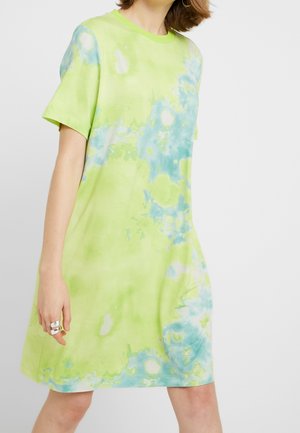 Kobieta w krótkim rękawie limonkowo-zielonej i niebieskiej luźnej sukience tie-dye, trzymająca rękę z srebrnymi pierścionkami, na jednolitym jasnym tle.