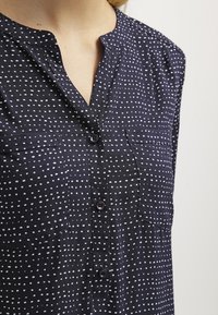 Camicia blu navy con punti bianchi, dotata di colletto e due tasche sul petto. Il tessuto sembra liscio con una leggera lucentezza.