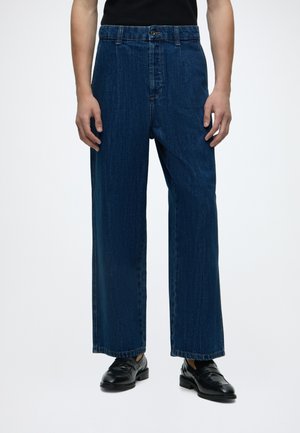 Person iført mørkeblå jeans med vidde ben og sorte læder loafers, stående mod en ensfarvet lys baggrund.