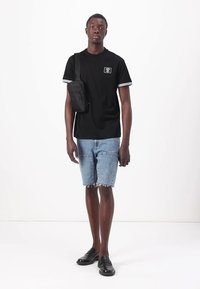 T-shirt preta de manga curta com detalhes brancos, combinada com calções de ganga desgastados e ténis pretos sem atacadores. A modelo leva uma pequena bolsa preta.