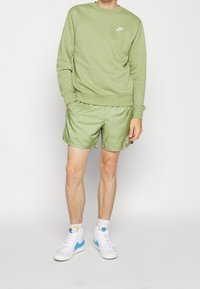 Ljusgrön sweatshirt med rund hals och Nike-logotyp, kombinerad med ljusgröna shorts. Vita höga sneakers med blå detaljer.
