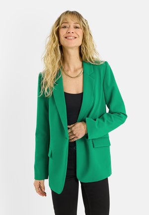 Femme souriante aux longs cheveux blonds portant un blazer vert vif par-dessus un haut noir et un pantalon noir, debout devant un fond blanc.