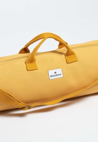 Bolso de lona amarillo con asas dobles y una correa para el hombro. Cuenta con un cierre de cremallera y un parche con el logo. Textura suave, diseño sencillo.