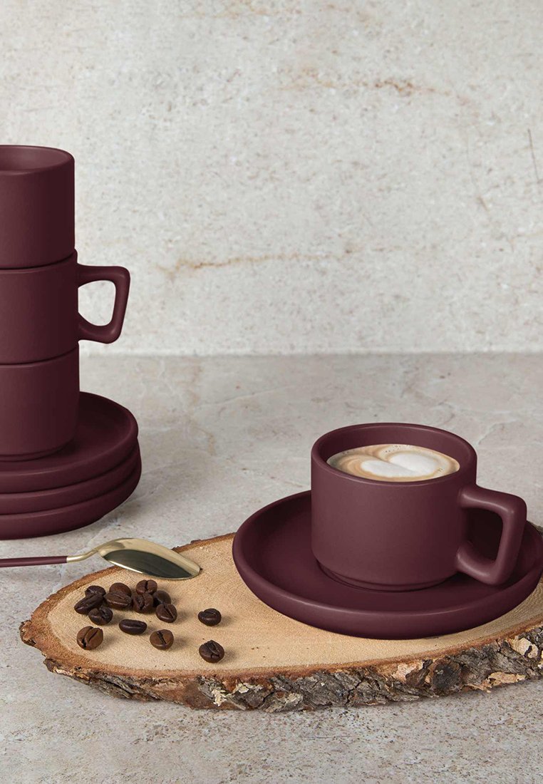 Taza de café de cerámica color malva con plato, tazas apiladas, cuchara de plata y granos de café sobre un tronco de madera. Textura suave, forma minimalista.