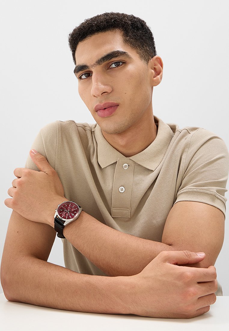 Ronde klok met een bordeauxrood vlak en zilveren accenten, zwartleren band, gedragen om een lichtgehuwde pols. Neutrale, beige poloshirt.