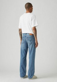 Jeans de mezclilla azul con corte recto, que presentan un parche de cuero marrón en la cintura trasera. Combinados con una camiseta blanca de manga corta y lisa.
