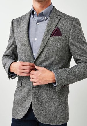 Hombre abrochándose una chaqueta de lana gris sobre una camisa azul clara, con un pañuelo de bolsillo púrpura oscuro, sobre un fondo claro y liso.