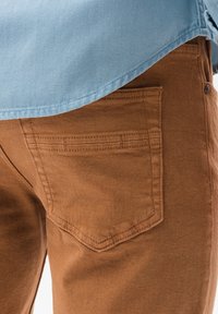 Pantalones de algodón marrón con un detalle de bolsillo trasero, combinados con una camisa de color azul claro cuya parte inferior es visible por encima. La tela tiene un acabado suave.