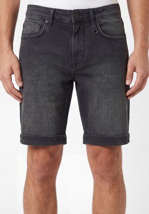 Pantalones cortos vaqueros negros de hombre con dobladillos remangados y bolsillos delanteros, usados por una persona de pie con las manos relajadas a los lados.
