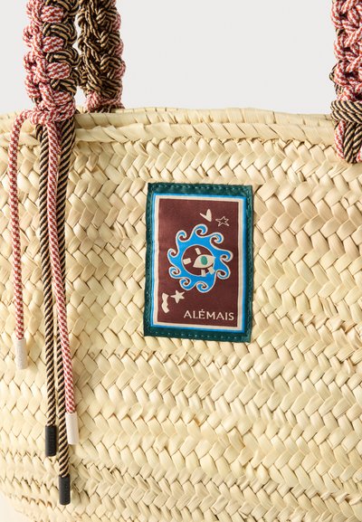 Πλεγμένη τσάντα από άχυρο με πλεγμένες ροζ και μαύρες λαβές και ένα πατς με τυρκουάζ περίγραμμα που φέρει ένα μπλε στροβιλώδες σχέδιο και κείμενο "ALÉMAIS."