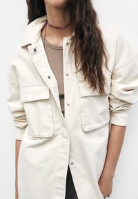 Beżowa koszula typu shirt jacket z zapięciem na guziki, dwoma przednimi kieszeniami i podwiniętymi rękawami. Noszona na dopasowanej brązowej odzieży, z koralikami na naszyjniku.