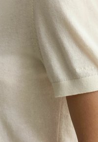 Hellbeige, kurzärmliger Pullover aus weichem Strickstoff. Verfügt über ein geripptes Bündchen mit einem dezenten Logo-Detail in der Nähe der Schulter.