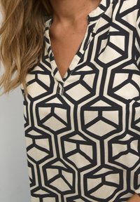 Femme portant un blouse à motif géométrique noir et blanc avec un col en V et des cheveux longs et ondulés sur un côté.