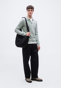 Νέος άνδρας που φοράει ανοιχτό πράσινο πουλόβερ, μαύρο φαρδύ παντελόνι, γκρι μπότες, κρατώντας μια μεγάλη μαύρη τσάντα ώμου, στέκεται μπροστά σε λευκό φόντο.