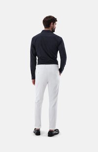 Mann, der ein schwarz-gestreiftes Hemd und weiße Hosen trägt, von hinten stehend. Schwarze Slipper und keine sichtbaren Muster auf den Hosen.