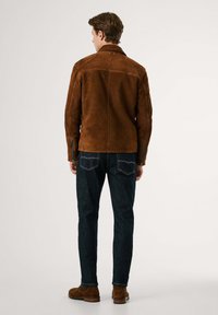 Giacca in suede marrone con taglio dritto, cuciture visibili e collare; abbinata a jeans in denim scuri e stivali in suede marrone.