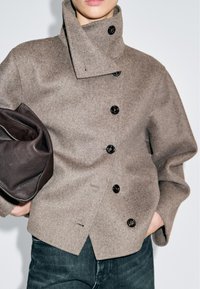 Giacca in lana marrone con colletto alto, dotata di bottoni neri e design corto. Abbinata a una borsa in pelle scura. Tessuto morbido.