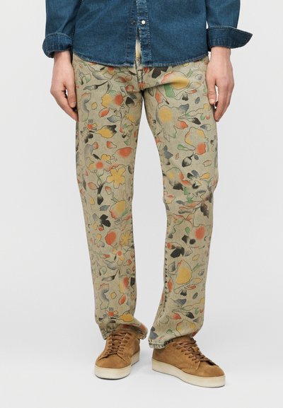 Hombre con pantalones beige con patrones florales abstractos multicolores, zapatillas de ante marrón y una camisa azul de mezclilla abotonada.