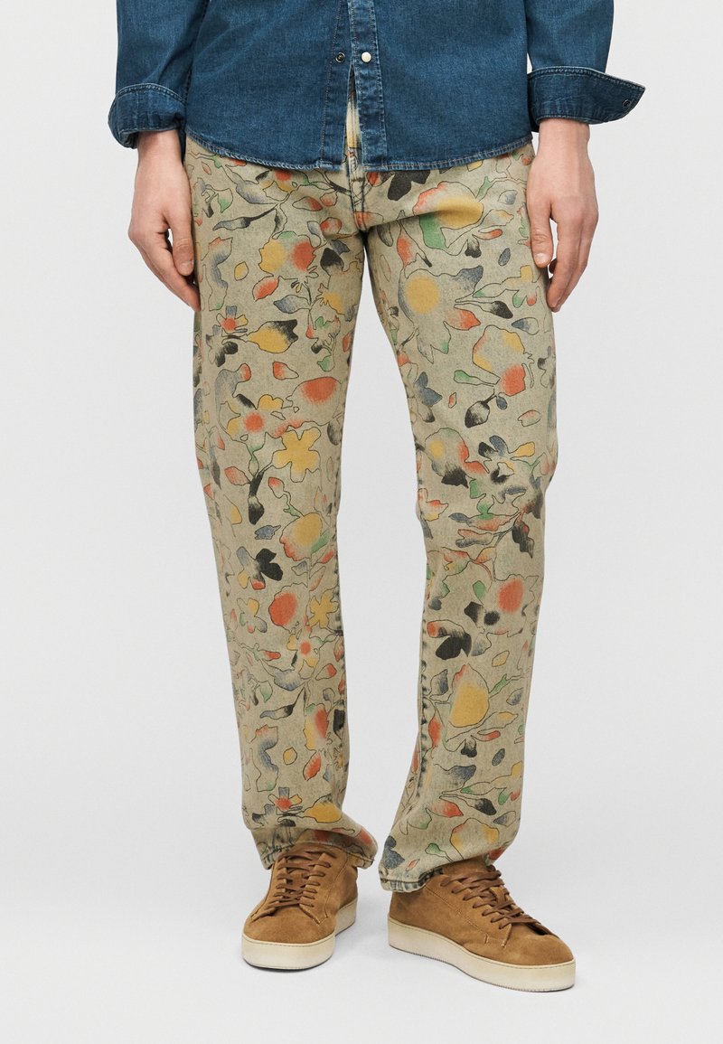Homme portant un pantalon beige avec des motifs floraux abstraits multicolores, des baskets en daim marron et une chemise en jean bleue boutonnée.