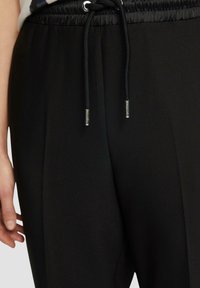 Czarne spodnie z dopasowanym pasem, elastyczną sznurówką oraz srebrnymi metalowymi końcówkami. Tkanina wydaje się gładka z detalami bocznych plis.