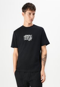 Black cotton t-shirt with short sleeves and a round neck, featuring a white graphic logo on the front. Model wearing black pants.