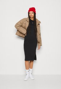 Brun pufferjacka, svart åtsittande klänning, vita sneakers och röd mössa. Modellen visar en avslappnad streetwear-stil med lager av texturer.