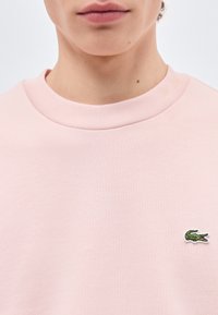 Κοντινό πλάνο ενός ατόμου που φοράει ροζ ανοιχτό πουκάμισο με μικρό πράσινο λογότυπο κροκοδείλου στο στήθος, εμφανίζοντας μόνο τα χείλη και τον λαιμό.