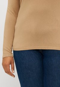Beige langermet topp med en jevn tekstur, tettsittende design og rund kant. Kombinert med mørkeblå denimjeans.