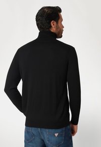 Maglione nero a collo alto con texture a coste, maniche lunghe e design aderente. Indossato con jeans blu con un patch del logo sulla tasca posteriore.