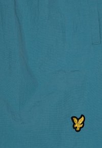 Lengvi turkio šortai su lygia tekstūra, ant kairės apatinės dalies esantis juodu kraštu apibrėžtas geltonas erelio emblema.